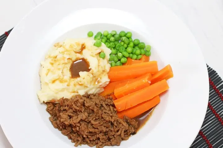 Seasoned ground beef with carrots, peas, potatoes and gravy on a white plate.